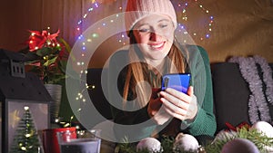 Young happy woman sitting on the sofa in Christmas decorated living room using mobile phone.