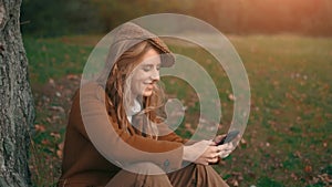 The young happy woman is smiling and touching a cell phone next to tree in a park or forest. A girl is chatting a text