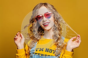 Young happy woman in sunglasses on bright yellow background. Blonde curly hair, cute smile