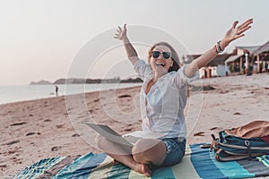 Young happy woman in sunglasses sitting on the beach with laptop, freelancer with open arms, remote work concept