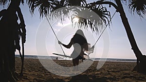 Young happy woman in swimsuit and shirt relaxing at swing at tropical ocean beach. Beautiful girl sitting on swing and