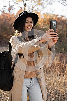 Young happy woman takes selfie photo during hiking in park, walking by autumn forest