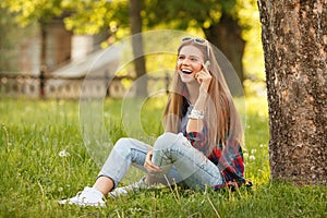 Young happy woman talking on cell phone sitting on grass in summer city park. Beautiful modern girl in sunglasses with a