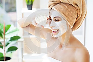 Young happy woman in towel, applying facial clay Mask in stylish bathroom.