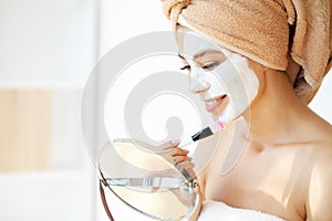 Young happy woman in towel, applying facial clay Mask in stylish bathroom.