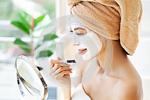 Young happy woman in towel, applying facial clay Mask in stylish bathroom.