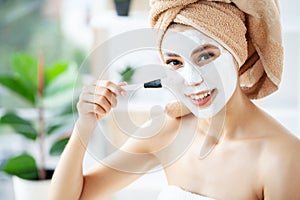 Young happy woman in towel, applying facial clay Mask in stylish bathroom.