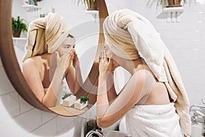 Young happy woman in towel applying organic face scrub and looking at round mirror in stylish bathroom. Girl making facial massage