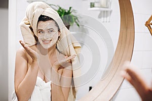 Young happy woman in towel applying organic face scrub and looking at round mirror in stylish bathroom. Girl making facial massage