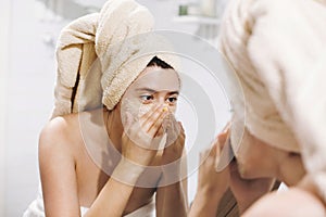 Young happy woman in towel making facial massage with organic face scrub and looking at mirror in stylish bathroom. Girl applying