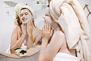 Young happy woman in towel making facial massage with organic face scrub and looking at mirror in stylish bathroom. Girl applying