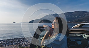 Young happy woman traveler enjoying the sunset at the sea while standing next to the car. Summer holidays and travel