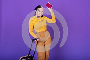 Young happy woman traveler with suitcase while showing her passport against purple background. Copy space