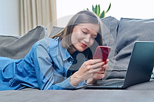 Young happy woman using smartphone laptop, lying on sofa at home