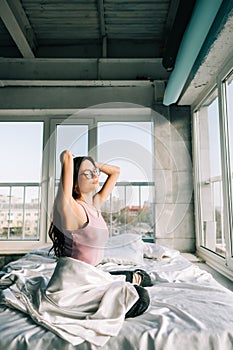 Young happy woman wake up in the morning, sitting on bed and lazily stretches hands