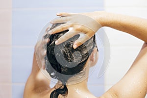 Young happy woman washing her hair with shampoo, hands with foam closeup. Back of beautiful brunette girl taking shower and