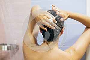 Young happy woman washing her hair with shampoo, hands with foam closeup. Back of beautiful brunette girl taking shower and