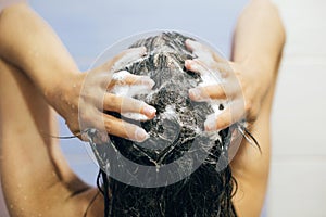 Young happy woman washing her hair with shampoo, hands with foam closeup. Back of beautiful brunette girl taking shower and