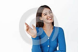 Young happy woman waving hand, greeting you, saying hello and smiling friendly at camera, standing in blue blouse over