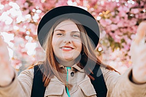 Young happy woman wearing hat taking selfie on spring city street. 30s girl smiling enjoying spring summer day. High