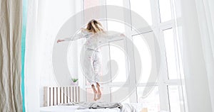 Young, happy woman wearing pajamas dancing, jumping on bed in her bedroom at home.
