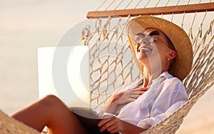Young happy woman wearing in straw hat and sunglasses enjoying freelance work on laptop while lying in the hammock on the beach