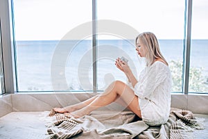Young happy woman in a white knitted dress enjoying sea view next to big window with cup of tea. Panoramic window. Floor coverings