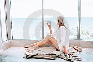 Young happy woman in a white knitted dress enjoying sea view next to big window with cup of tea. Panoramic window. Floor covering