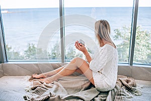 Young happy woman in a white knitted dress enjoying sea view next to big window with cup of tea. Panoramic window. Floor coverings