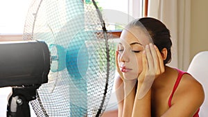 Young headache woman suffering a heat wave and using a fan sitting on a couch in the living room at home