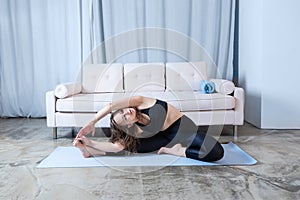 Young healthy beautiful woman in sports top and leggings practicing yoga at home, sitting in pose on yoga mat. side body stretch