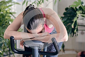 Young healthy fit woman training at home on exercise bike during work-out feeling exhausted and dizzy, lowered his head on his han