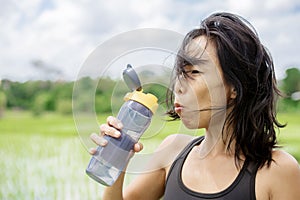 Young healthy sporty Asian Chinese woman drinking water bottle after fitness training and running workout outdoors on green field