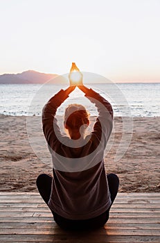 Young healthy woman practicing yoga holding hands in meditation pose with sun between themon the beach at sunrise