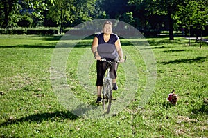 Young healthy woman rides bike in park with small dog running near.