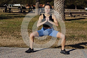 Young healthy woman in sportswear listens music in headphones while doing fitness squat leg exercises outdoors in park