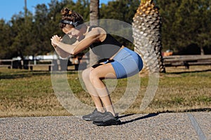 Young healthy woman in sportswear listens music in headphones while doing fitness squat leg exercises outdoors in park