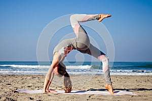Young healthy woman in a stylish one-piece jumpsuit practicing yoga on the beach at sunrise