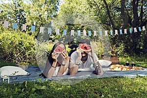 Young heterosexual couple laying on the ground on a blanket having picnic and looking through piece of watermelon