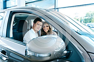 Young heterosexual couple sitting in a car in showroom and smiling through the open