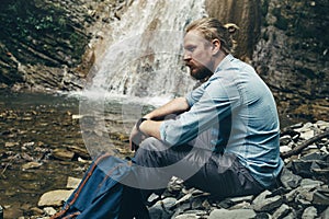 Young Hiker Man With Backpack, Sitting On The Rocks Near Waterfall Trek Hiking Destination Experience Lifestyle Concept