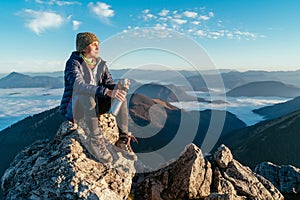 Young hiker woman sitting on the mountain summit cliff, drinking tea from a thermos flask and enjoying mountains valley covered
