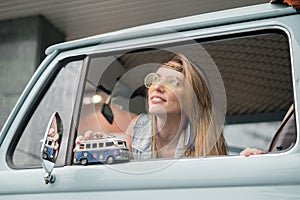 Young hippie woman driving a classic van with a toy car model in her hand. Smiling blurred girl holding a miniature