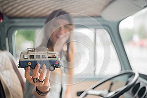 Young hippie woman driving a classic van with a toy car model in her hand. Smiling blurred girl holding a miniature