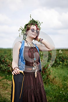 Young hippie woman with short red hair, wearing boho style clothes, sunglasses and flower wreath, standing on green field, holding