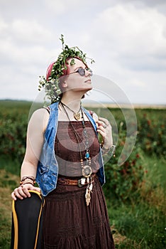 Young hippie woman with short red hair, wearing boho style clothes, sunglasses and flower wreath, standing on green field, holding