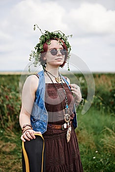 Young hippie woman with short red hair, wearing boho style clothes, sunglasses and flower wreath, standing on green field, holding