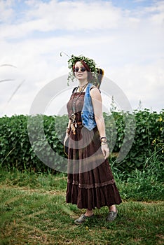 Young hippie woman with short red hair, wearing boho style clothes, sunglasses and flower wreath, standing on green field, holding