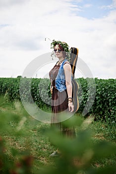 Young hippie woman with short red hair, wearing boho style clothes, sunglasses and flower wreath, standing on green field, holding