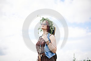 Young hippie woman with short red hair, wearing boho style clothes, sunglasses and flower wreath, standing on green field, holding